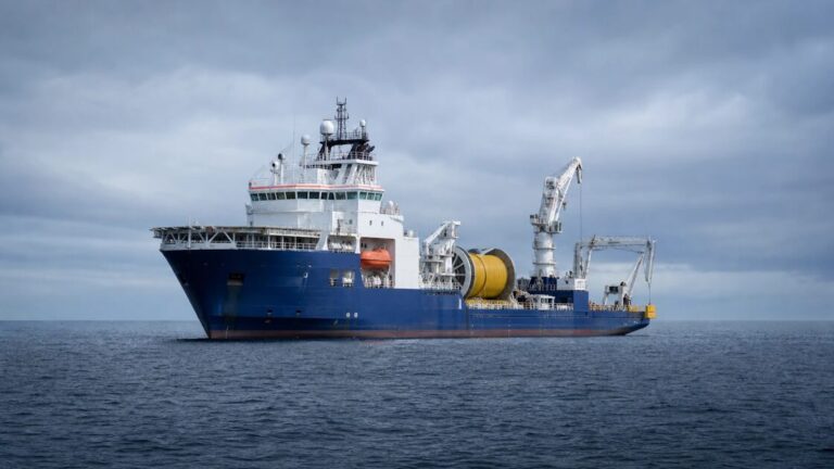 Large white ship with a bulky stern winch on open gray sea, laying cable from a spool.